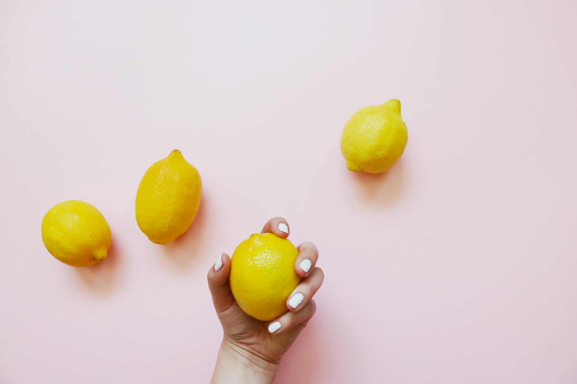 A hand holding a lemon on a soft pink background, surrounded by additional lemons.