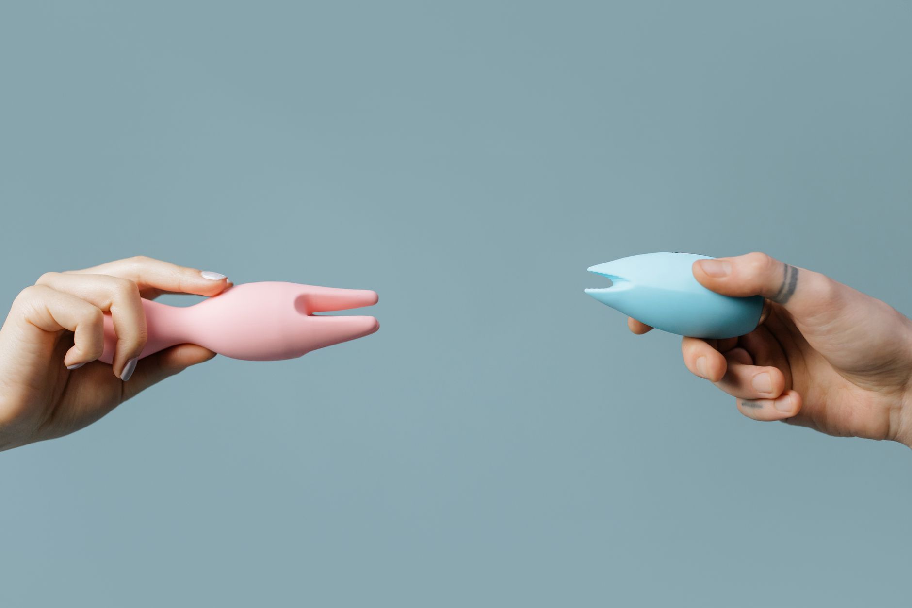 Two hands holding silicone vibrators against a pastel background.